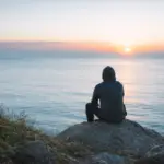 Person sitting quietly on a coastal rock at sunrise illustrating why the brain needs quiet and mental clarity